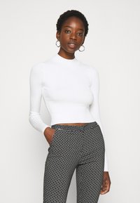 Haut à col montant blanc à manches longues et côtelé, associé à un pantalon tailleur noir et blanc à motifs, avec poches latérales et une taille lisse.
