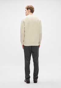 Ανοιχτό μπεζ ριχτό πουλόβερ με μακριά μανίκια, στρογγυλή λαιμόκοψη και άνετη εφαρμογή, συνδυασμένο με σκούρες γκρι παντελόνες και μαύρα παπούτσια.