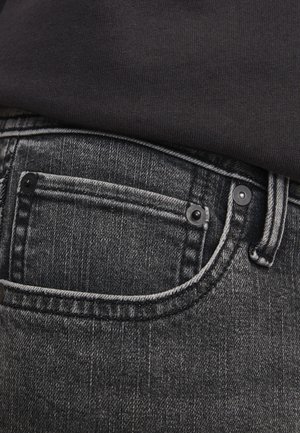 Mørkegrå denimbukser med en lomme på toppen, kontrastsøm og metalnitter. Teksten ser litt slitt ut med vertikale linjer.