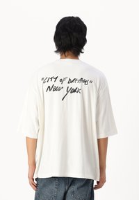 Hvid oversized T-shirt med korte ærmer. Bagsiden prydes af sort håndskrevet tekst: "CITY OF DREAMS" og "New York." Blødt bomuldsstof.