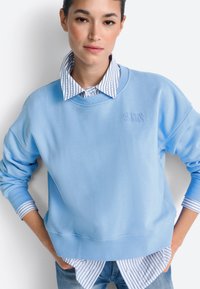 Hele sinine dressipluus, mis on kihiliselt kantud sinise ja valge triibulise kraega särgi peale. Pehme kangas koos soonikkoes mansettide ja tikitud logoga.