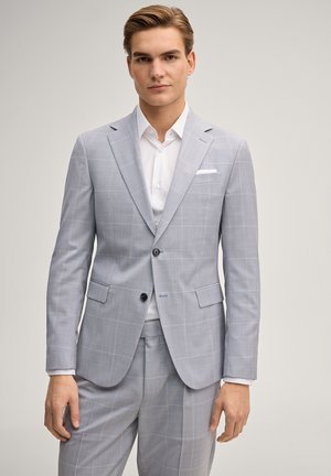 Giovane uomo con indosso un abito a quadri grigio chiaro e una camicia bianca elegante, in piedi con un'espressione neutra su uno sfondo semplice.