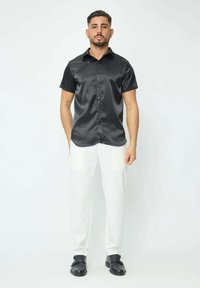 Chemise noire en satin à manches courtes avec un bouton sur le devant, associée à un pantalon blanc et des chaussures noires, se tenant devant un fond blanc uni.