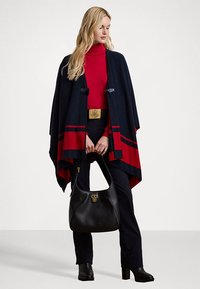 Poncho navy dan merah dengan desain geometris, atasan turtleneck, celana hitam ramping, dan tas jinjing hitam dengan perkakas emas.