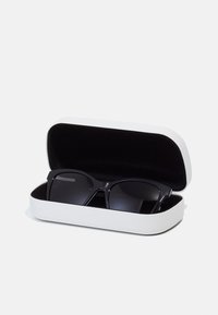 Ochelari de soare negri cu un cadru dreptunghiular într-un etui alb și negru. Etuiul are un exterior neted și o căptușeală interioară moale.