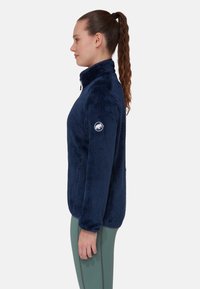 Giacca in pile blu navy con colletto alto, superficie tessurizzata, zip laterale e patch del logo sulla manica sinistra. Design aderente.