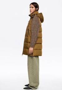 Chaleco acolchado color caqui con capucha, bolsillos con cremallera y herrajes plateados, combinado con un suéter marrón texturizado y pantalones de pierna ancha color oliva.