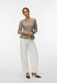 Femme portant un pull gris à manches longues, un pantalon blanc à jambes larges, et des chaussures à talons marron, debout les mains jointes devant elle.