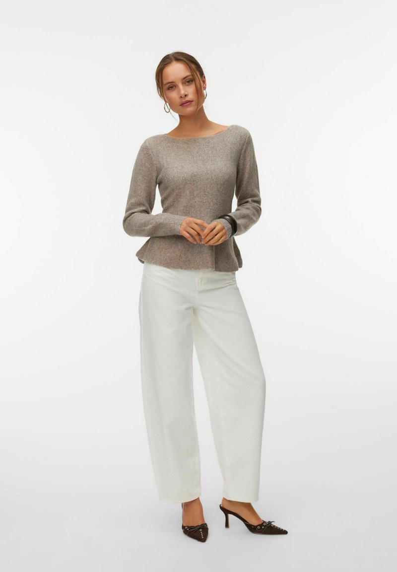 Femme portant un pull gris à manches longues, un pantalon blanc à jambes larges, et des chaussures à talons marron, debout les mains jointes devant elle.