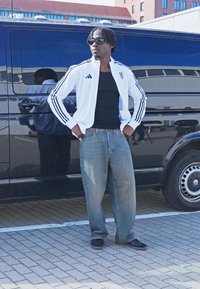 Hvid Adidas træningsjakke med sorte striber, sort tanktop, løse blå jeans og sorte sko, stående ved siden af en varevogn på asfalt.