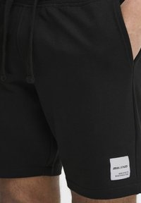 Pantaloni corti neri in cotone con vita regolabile con cordino, tasche laterali e un'etichetta bianca con la scritta "BLEND" e testo. Tessuto liscio e design casual.