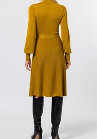 Senapsgul, knälång klänning med en figurnära bodice, långa puffärmar och texturerad ribbning, kombinerad med svarta knähöga stövlar.