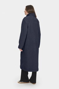 Marinblå lång kappa i texturerad fleece med hög krage och avslappnad passform, tillsammans med svarta byxor och beige skor.