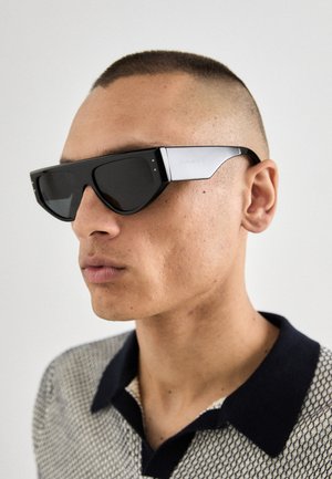 Occhiali da sole neri con montatura angolare, dettagli in argento e lenti scure. Il modello indossa una maglietta in maglia texturizzata con colletto blu navy.