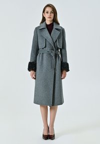 Cappotto in lana grigia con colletto, vita con cintura e polsini in pelliccia sintetica. Design fino al ginocchio, caratterizzato da una texture morbida e una silhouette classica.