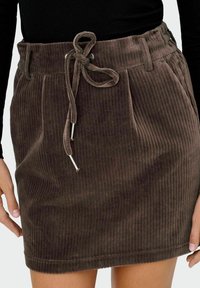 Gonna di velluto a coste marrone con vita regolabile a coulisse indossata con una maglia nera a maniche lunghe, con le mani ai lati.