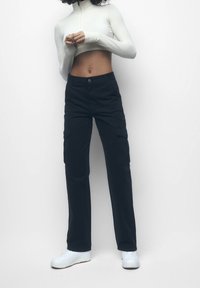 Pantalons cargo noirs avec une coupe décontractée, présentant des poches latérales et une fermeture par bouton, associés à un haut court à manches longues blanc et des baskets blanches.