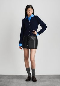 Marineblaue Strickjacke mit goldenen Knöpfen, kombiniert mit einem schwarzen Leder-Minisrock mit Knopfleiste vorn, blauem Hemd, grauen Socken und dunklen Loafers.