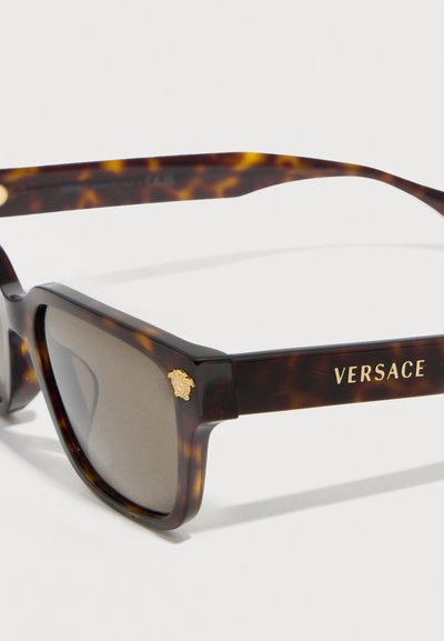 Γυαλιά ηλίου Versace με σχέδιο χελώνας, ορθογώνιοι φακοί καφέ χρώματος, χρυσό έμβλημα Μέδουσας στη γωνία και λογότυπο Versace στο μπράτσο του σκελετού.