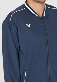 Marineblå zip-up jakke med hvide accenter, ribbestrikket krave, letvægtsstof og logo på venstre bryst. Har sidelommer.