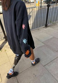 Czarna, oversize bluza z wielokolorową haftowaną dekoracją kwiatową na rękawach. Czarnie skarpetki z wzorem oraz widoczna proteza nogi.