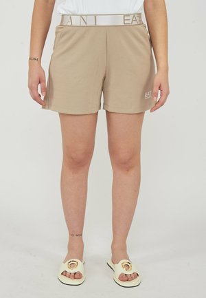 Persoon draagt beige EA7-short met elastische logoband en crèmekleurige slidesandalen met gouden ringdetail, staand tegen een effen achtergrond.