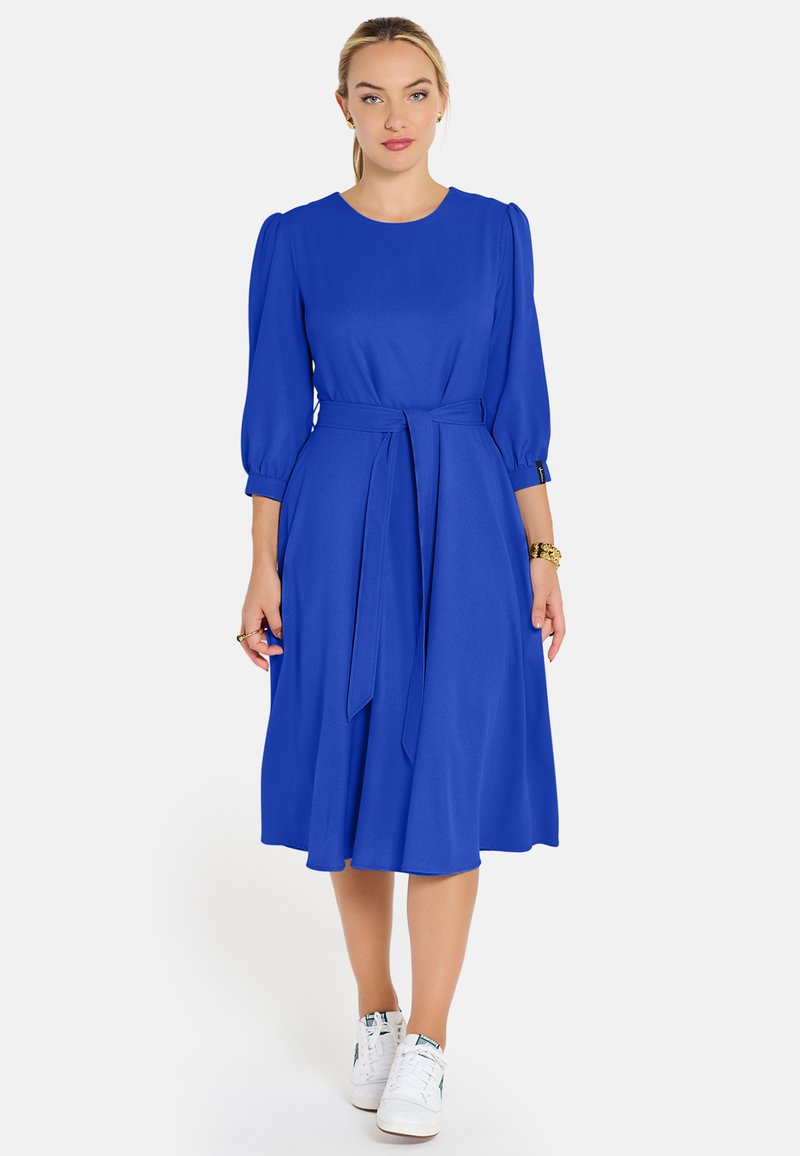 Žena v kráľovsky modrej midi šatách s nariasenými trojštvrťovými rukávmi, s opaskom v páse a bielymi teniskami, stojaca na neutrálnom pozadí.
