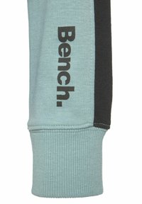 Helles blaues Sweatshirtmaterial mit einem schwarzen Seitenpanel, das das Wort "Bench." in fetter, schwarzer Schrift entlang des Ärmels trägt. Gerippte Bündchen.