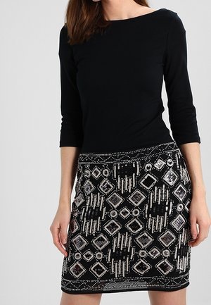 Femme portant un haut noir à manches trois-quarts et une jupe noire avec des motifs géométriques blancs et perles.