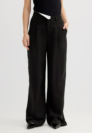 Persona que lleva pantalones negros sueltos de cintura alta con bolsillos, combinados con un top negro sin mangas y zapatos negros puntiagudos.