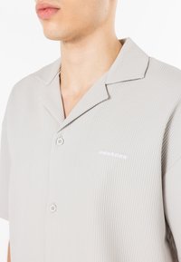 Camisa de manga curta de riscas cinza-claro com colarinho notched, apresentando dois botões e um pequeno logótipo branco no painel esquerdo do peito.