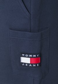 Námořnická mikina s texturovaným povrchem, má přední kapsu a obdélníkovou nášivku s logem "TOMMY JEANS" v bílé, červené a modré barvě.
