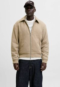 Chaqueta beige con cremallera hecha de un tejido texturizado, que presenta un cuello con solapa, puños acanalados y dos bolsillos frontales sobre un suéter blanco.