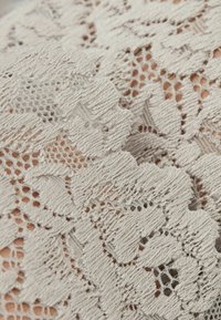 Tela de encaje en beige claro con patrones florales. Presenta un acabado texturizado y bordes de picos, que revelan intrincados detalles de costura.