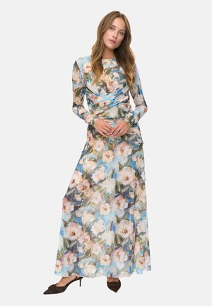 Femme portant une robe florale à manches longues, traînante, dans des teintes pastel de bleu, rose et vert, associée à des talons aiguilles noirs, se tenant avec les mains jointes.