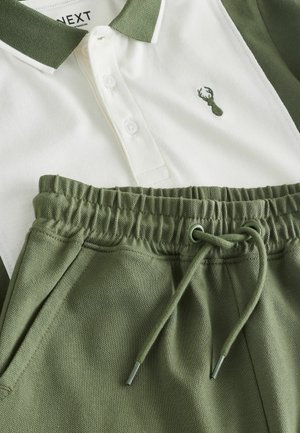 Zelena i bijela polo majica s ovratnikom i zatvaračem na gumbe, s izvezenim logotipom jelena. Uz nju se nose zelene kratke hlače s elastičnim strukom.