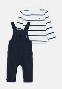 Fehér, hosszú ujjú ing sötétkék csíkokkal és kis logóval, amelyet sötétkék overallal párosítottak, állítható pántokkal és elülső zsebből.