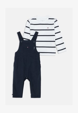 Balta ilgomis ranksovėmis marškiniai su tamsiai mėlynomis juostelėmis ir mažomis logotipu, derinami su tamsiai mėlynomis kombinezoninėmis kelnėmis, turinčiomis reguliuojamus diržus ir priekinę kišenę.