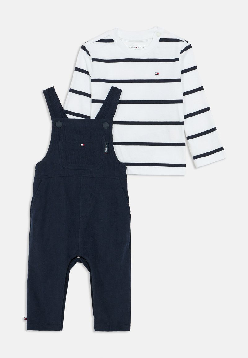 Fehér, hosszú ujjú ing sötétkék csíkokkal és kis logóval, amelyet sötétkék overallal párosítottak, állítható pántokkal és elülső zsebből.