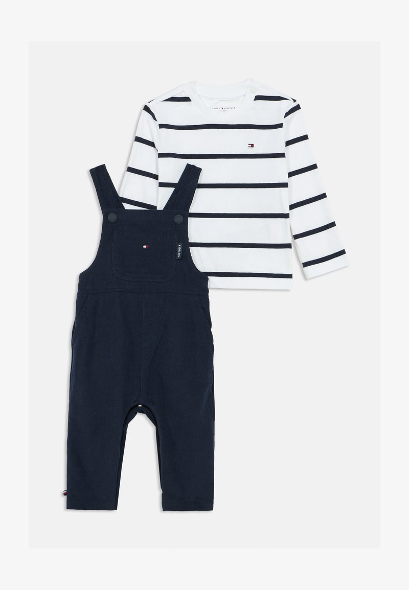 Λευκή μακρυμάνικη πουκάμισο με ναυτικές ρίγες και μικρό λογότυπο, συνδυασμένη με ναυτικές φόρμες που διαθέτουν ρυθμιζόμενα λουριά και μπροστινή τσέπη.