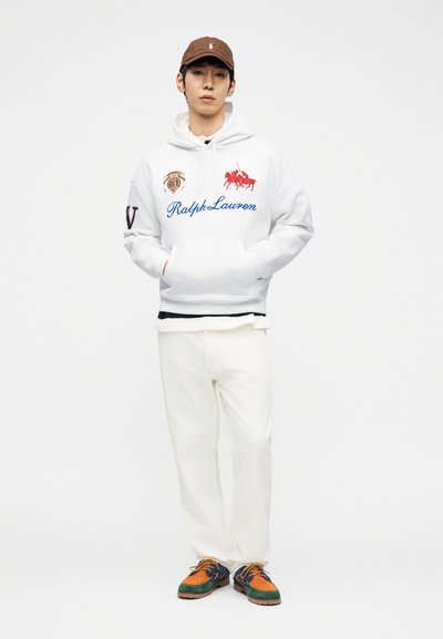 Νέος άνδρας που φοράει λευκή μπλούζα με κουκούλα Ralph Lauren, λευκό παντελόνι, καφέ καπέλο και πολύχρωμα casual παπούτσια, στέκεται με τα χέρια στις τσέπες.