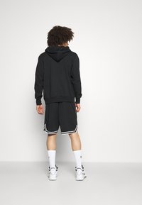 Sudadera negra con capucha ajustable, combinada con pantalones cortos negros con detalles en blanco. El conjunto se completa con calcetas blancas y zapatillas deportivas.