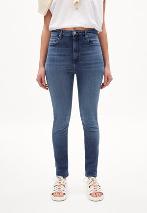 Personne portant un jean bleu taille haute slim, un t-shirt court blanc et des sandales blanches à lacets, debout devant un fond uni.
