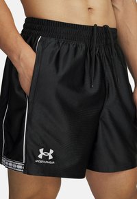 Črne športne kratke hlače z elastičnim pasom in vrvico, ki imajo bele stranske črte in logotip Under Armour na robu.