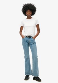 Camiseta blanca de manga corta, jeans acampanados de color azul claro con una textura desteñida y calzado negro. El modelo está de pie con las manos en las caderas frente a un fondo blanco.