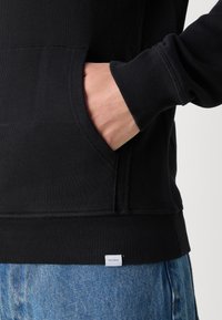 Czarna bluza z kieszenią, ściągaczami przy rękawach i u dołu. Widoczna metka marki "LES DEUX" na dolnym wykończeniu. Zestawiona z niebieskimi dżinsami.