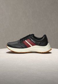Baskets en cuir noir avec des bandes latérales blanches et bordeaux, semelle épaisse blanche, semelle extérieure en gomme, et logo "BALLY" sur le panneau latéral.