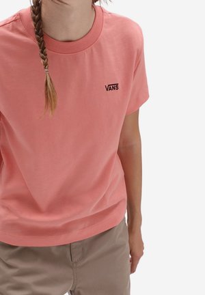 Persona con cabello trenzado que lleva una camiseta rosa de Vans y pantalones beige sobre un fondo liso.