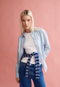 Lichtblauw knoopshirt over een witte getailleerde t-shirt, met een blauw-wit gestreept truitje om de taille, gecombineerd met blauwe jeans.