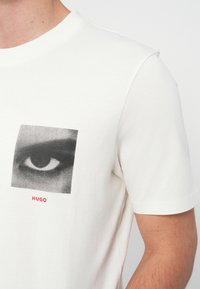 Biała koszulka z czarno-białym zbliżeniem oka i czerwonym napisem "HUGO" poniżej, na wysokości klatki piersiowej.